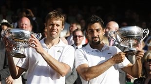 Daniel Nestor left, and Nenad Zimonjic of Serbia hold up their trophies, after defeating Bob and Mike and Bryan of the US in the men's doubles final on the Centre Court at Wimbledon in 2009.  Both men are in white. Each is holding a large silver-bowl trophy. Nestor, blond with short hair, contrasts with Simonjic's who has dark hair and a goatee. Neither is really smiling but offering somewhat half-smiles.
