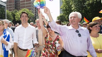 Liberal leader Justin Trudeau, left, and Bloc Quebecois Leader Gilles Duceppe wave to supporters during Sunday's annual gay pride parade in Montreal, Sunday. NDP leader Tom Mulcair and Green Party leader Elizabeth May also attended while Stephen Harper campaigned in Ottawa. Duceppe, who is developing a paunch, wears a blue dress shirt with the sleeves rolled up just a tad. He holds gay pride flag high with his right hand. In his left hand is a small plastic water bottle. He wears dark pants and has a pair of sun glasses tucked into the top of his shirt. Trudeau in a white shirt and brown pants, is looking into the distance. If it were theatre, he would be playing the gods.