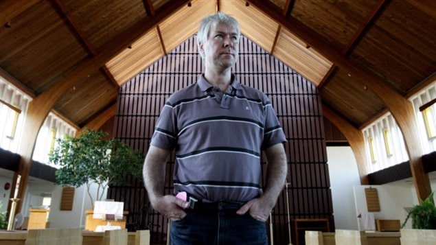 Former KGB agent Mikhail Lennikov in a 2009 photo at Vancouver's First Lutheran Church, shortly after he sought sanctuary. We see a man wearing a grey and black polo shirt standing with his hands in the front pockets of his blue jeans. His face under short grey hair wears a look of chagrin. Behind and above him, we see a modern church constructed mainly of dark wood. The apex of the roof and ceiling rises above him.