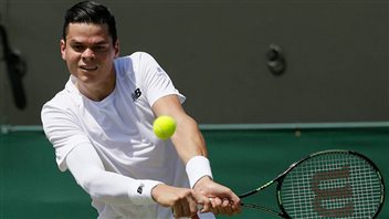 Milos Raonic has struggled since Wimbledon, is the No. 10 seed in New York. Raonic is about to hit a two-handed backhand. We see the yellow ball approaching. He is dressed all in white with an elastic white sleeve on his right arm and white wrist bands on both wrists. A bear of a man, his hear is jet black and slicked back.