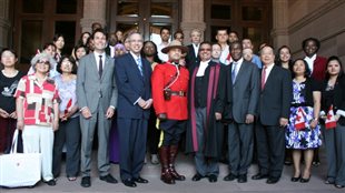 New Canadians have to take the Oath of Citizenship as one of the final steps before becoming a citizen. We see a group of new Canadians (about forty) of many nationalities gathered on steps in front of a courthouse. In the front row, a Mountie in full dress uniform stands next to robed judge. Virtually all in the picture have wide smiles on their faces.