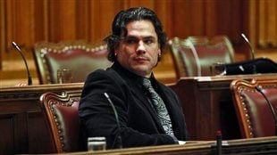 Patrick Brazeau was appointed to the Senate by Prime Minister Harper in 2008. We see Brazeau seated in the Senate wearing a dark jacket and fancy tie. A handsome man with long, black hair, he has his right arm resting on the desk in front of him as he looks to his right.