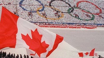 The opening of the 1988 Winter Olympic Games in Calgary. Is the city ready for an encore presentation? We see a giant Canadian flag in the foreground. Below the flag, athletes are marching into the stadium. In the background are fans holding cards to form the five Olympic rings.