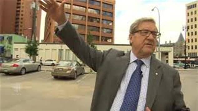 Lloyd Axworthy was Canada's minister of foreign affairs from 1996 until 2000. We see Axworthy in a grey suit and dark blue tie on a street on what appears to be a dank autumn day with his right hand raised, apparently hailing a taxi. His grey hair is well-groomed and wears dark-rimmed glasses.