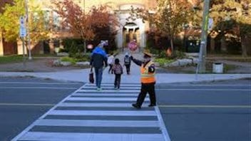 For students across Canada, September is the real start of a new year. We see an elderly male crossing guard in an orange vest and dark pants stopping traffic is his right hand as a mother and her tiny child hold hands as they head toward the entrance of a school that is surrounded by large trees bathed in fall colours.