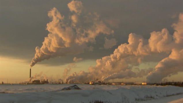The Quebec government wants to cut emissions levels considerably by 2030. We look across a large field of snow towards a series low-slung factory smokestacks  spewing large, cumulus-like clouds of smoke skyward towards the right of the photo. The sky appears ominously dark.