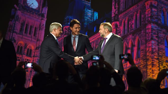 Conservative leader Stephen Harper, left, greets NDP leader Tom Muclair as Liberal leader Justin Trudeau looks on Thursday night in Calgary. All are dressed in sharp suits. Both Harper and Muclair are greying. Trudeau looks as if he could be their son. Behind them bathed in red and blue lighting we see a giant photo of the Peace Tower and Parliament Hill.