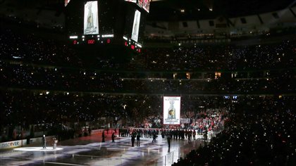 The Chicago Blackhawks' Stanley Cup championship banner is raised to the United Center ceiling during a ceremony before their game with the New York Rangers on Wednesday. The stands in the arena are aglow with cellphone lights reaching to the upper rafters. The ice blow reflects the flood lights (in streaks). At the far end, we see a lot of people is suits as the giant white banner makes its way up.