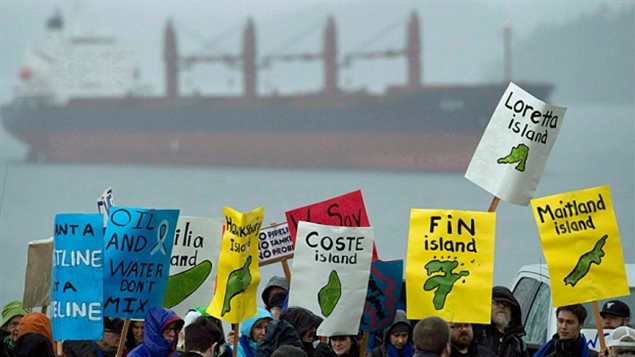 The proposed Northern Gateway pipeline has been a divisive issue in B.C. In the lower foreground of the photo, we see a group of about 20 protesters. They are holding blue, yellow, white and green placards, on which are written "Oil and Water Don't Mix", "Coste Island", "Fin Island", "Maitland Island" and "Loretta Island." Under the words on the signs are drawings of the individual islands. In the background across a small expanse of water, we see a large four-smoke stack freighter, painted orange and black.