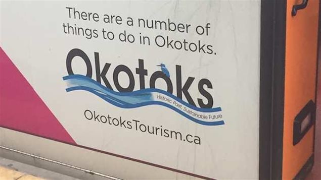 The slogan that launched a thousand quips. We see the side of some sort of a mainly white rectangle box, coloured orange at the right end with a red triangle shape in red covering the bottom of the left front. On the side facing us is the slogan, "There are a number of things to do in Okotoks."  Below those words, printed in bold-face, is the world "Okotoks" with what appears to be a tiny blue jay sitting atop the third "o." The word appears to be floating on a blue ribbon, in which the words "Historic Past, Sustainable Future" are written. Below the logo, is written "Okotoks Tourism.ca."