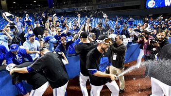 Toronto Blue Jays players were no pikers when it came to sharing their victory champagne on Wednesday. We see four players in their dark and soaked victory tee-shirts with bottles, shooting spay in all directions. They are at the edge of the lower stands where a group of fans, dressed mainly in Blue Jay blue, appear very happy to partake.
