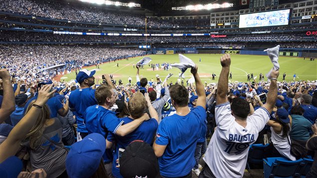 Toronto fans were ecstatic after the Wednesday's victory. In the seventh inning, not so much. We have a long shot from the back of the lower deck between home and first. Fans, many of them dressed in blue and white Jays jerseys and tee-shirts, have their arms raised in celebration, many of them hugging each other. In the distance gathered around the pitcher's mound at the centre of the green artificial turf, some winning Jays have gathered to celebrate as their teammates stream from the dugout the join them. We have a wide, circular shot of Rogers Centre, absolutely crammed with fans stands lit by the big overhead lights and the giant scoreboard. 