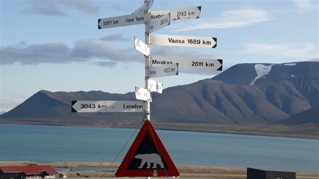 A signpost and a polar bear warning sign at Svalbard’s Longyearbyen airport is shown with blue waters and mountains in the background.