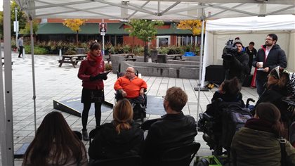 Valerie Burnet addresses supporters and members of the media on Saturday as Martin Beauregard looks on. Burnet is trim and stylish, with her dark hair pulled back. She wears a red scarf and a thigh-length skirt