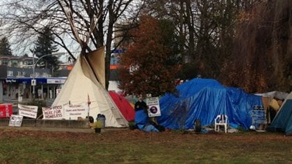 Residents at the homeless protest camp in Jubilee Park in Abbotsford, shown in 2013. To the left the photo we see a white tee-pee shaped tent. On the right, we see a smaller, formless blue tent. The day is overcast and the grass in front of the tents looks very moist.