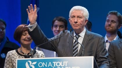 Bloc Québécois Leader Gilles Duceppe is getting out of politics. We see Mr. Duceppe in a grey suit and striped tie standing behind a podium waving with his right hand with pursed lips and a sad face. To his left, his wife, Yolande Brunelle, who had short, dark hair, stands. Her eyes are closed and she appears on the verge of tears. Behind the couple, several young men, likely family, appear to attempting not to show their sadness.