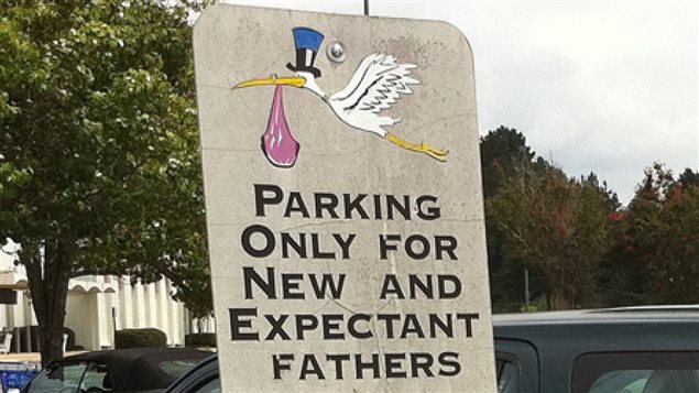 A new Canadian study indicates that not everything is always rosy for a lot of expectant fathers. We see a large grey sign in front of a parking lot. At the top of the sign is a drawing of our old friend, the baby stork. He is mainly white and carries a bundle from his yellow beak. On his head, he wears an blue, black and white Uncle Sam-shaped hat. Below the stork are written the words, "Parking Only for New and Expectant fathers." Behind the sign and cars, we see the dark green of trees, their leaves if full bloom. The sky is a light grey.