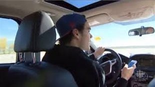 Driver distraction is a factor in four million vehicle crashes a year in Canada. We see from the back seat a young man wearing a black sweatshirt and blue baseball cap turned backward. His left hand in on the steering wheel of his car and his right hand holds a device that appears to be grabbing all his attention.