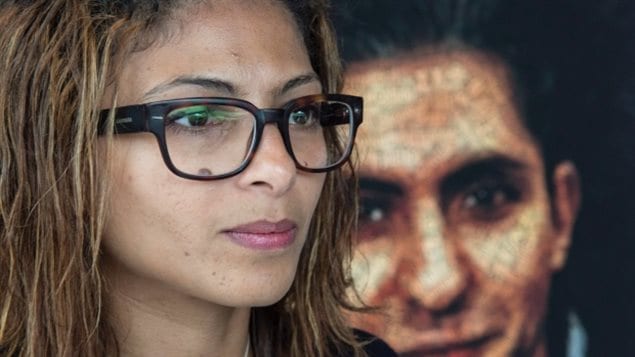  Ensaf Haidar's husband, Raif Badawi, is currently serving a 10-year prison sentence in Saudi Arabia. In the foreground on the left, we see Ms. Haidar. She has slightly stringy blond hair, dark eyes and wears large dark glasses and a determined look. Her lips are slightly pursed. Behind her over her left shoulder is a large photo of her husband. He has a dark complexion under black hair and kind, dark eyes.