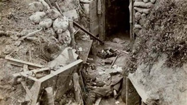 The body of a dead German soldier lies near the entrance to his dugout. While the entrance is intact, the sides of the trench have collapsed. Common items such as an entrenching tool, a water bottle, and ammunition pouches lie scattered on the ground