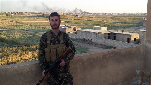   Photo of Canadian volunteer John Gallagher posted on his Facebook page on October 30, 2015. Gallagher, clad in camouflage fatigues and armed with an AK-47 assault rifle with ammunition pouches on his chest, stands on a balcony overlooking a rural area with smoke billowing in the distance.