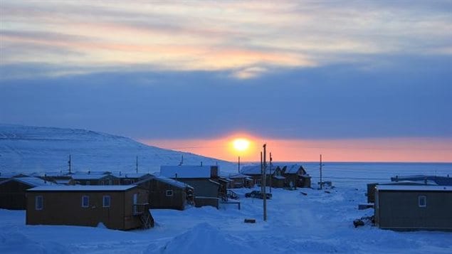 The Arctic village of Ulukhaktok, Canada is pictured covered with snow and with the sun setting behind it. 