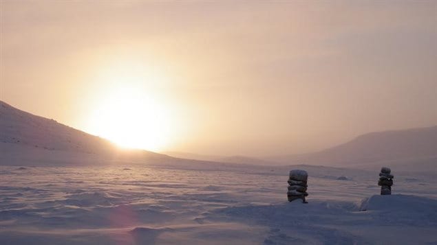 The sun sets on a snowy Arctic landscape with two small inukshuks in the foreground.