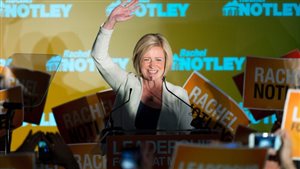Rachel Notley on the night of her stunning election victory in Alberta in May. Wearing a white sweater, she is waving with her left hand behind a microphone at a podium. Under her short blond hair is a radiant smile.