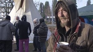 Abbotsford and won a major legal victory in 2015. We see an elderly man dressed in a parka in the foreground sipping coffee while five of his colleagues huddle outside a white teepee in the background.