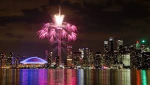 Opening Night at the Pan Am Games lit up downtown Toronto. We see downtown Toronto from across the water. Fireworks of all colours light up the sky and the city below, much of them reflecting in the calm of Lake Ontario.