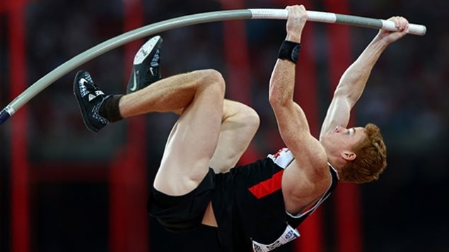 Onward and upward towards Rio, Shawn Barber won gold in the pole vault and the World Track and Field Championships in Beijing. We have a side view of Barber in mid-air. His long grey and white pole is bent toward him and his body is coiled as he attempts to make the jump.