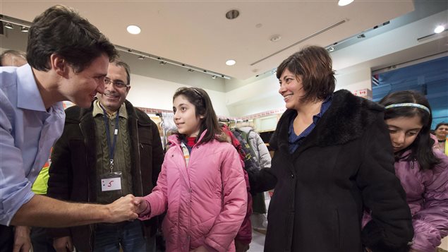 Prime Minister Trudeau greeted newly-arrived Syrian refugees at Pearson International airport in Toronto on Dec. 11. We see Mr. Trudeau, jacketless at the left of the picture, shaking the hand of a young girl dressed in a pink winter coat.  She stands between her father (to her right). who wears a light winter jacket, and her mother (to her left), who wears a full-length dark winter overcoat. A younger sister, who we barely see, has been hit by a bout of shyness and hides behind her mother. With the exception of the girl hit with shyness, everyone is smiling, though the girl in the centre does appear slightly bewildered.