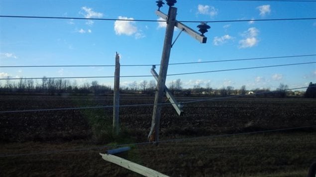 Blackouts caused by high winds are affecting thousands of hydro customers in Ontario and Quebec. We see a hydro pole standing at a 45 degree angle that has snapped off from its base, which remains planted in the ground. The countryside behind it is a dark brown colour while the sky with several scattered white clouds remains blue.