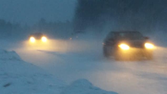 Coming--likely Monday night--to a road like this: winter chaos. We are looking at a virtual white-out. We see two cars through the white snow. They are travelling single file on a road surrounded by snow at its edge. It is evening so the cars' lights contribute to an eerie atmosphere.