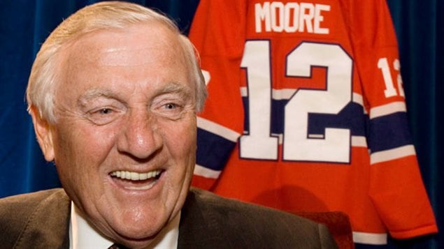 Dickie Moore wore the Canadiens' No. 12 with pride and played a key role on the teams many consider the greatest of all time. We see Moore at the left of the photo. He has a wide face, groomed, grey hair and a wide smile. Behind him his his hockey sweater bearing his name and the number 12.