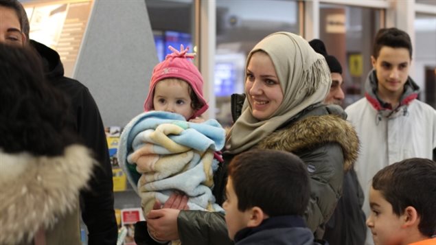 A mother and her child, refugees originally from Syria, wait inside the Saskatoon airport earlier this month before stepping into the chilly weather outside. We see a lovely woman dressed in a winter jacket with fur at the collar and a head scarf holding a baby wrapped in blanckets. Smiling, she is surrounded by two young boys to her left and a teen-aged boy behind her.