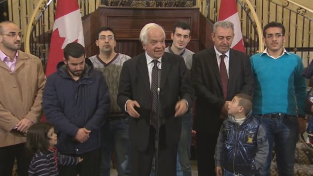 Immigration Minister John McCallum stands with a group of recently arrived Syrian refugees in Toronto in November. We see the white-haired and round-faced McCallum in a brown suit and tie gesticulating behind a thing microphone. He is surrounded by a group of men, mainly dressed in casual clothes. They are listening, as are two young children--a girl of about four or five on the left and a boy about about seven on the right. The girl is grabbing her father's jacket. The boy is looking up at McCallum with wide eyes.