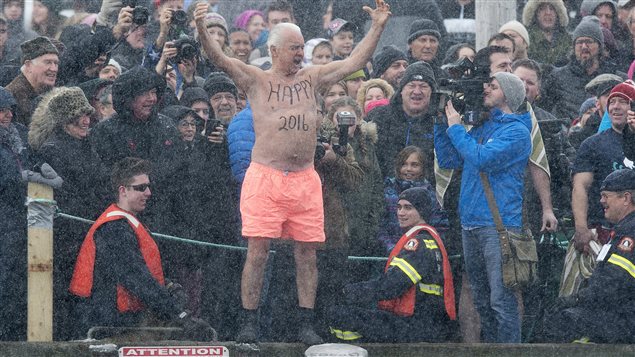 Ernie Ross, 81, prepares to jump into the North Atlantic in Herring Cove, N.S. on Friday. Ernie is standing at the top of a dock by the edge of the sea wearing what appears to be pink underwear or a salmon-coloured swimsuit. His hands are raised in the air in triumph. He is surrounded by spectators, some emergency personnel and a lone television cameraman.