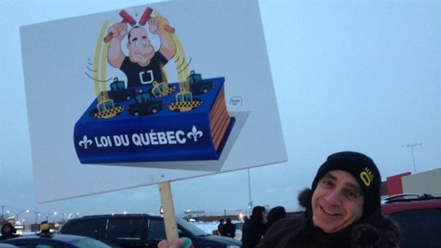 Elie Boujaoude was one of the taxi drivers taking part in Wednesday's protest. We see a young man in a toque at the right of picture. He is holding up a placard with a cartoon figure sitting on a blue law book on which is printed "Loi du Quebec."