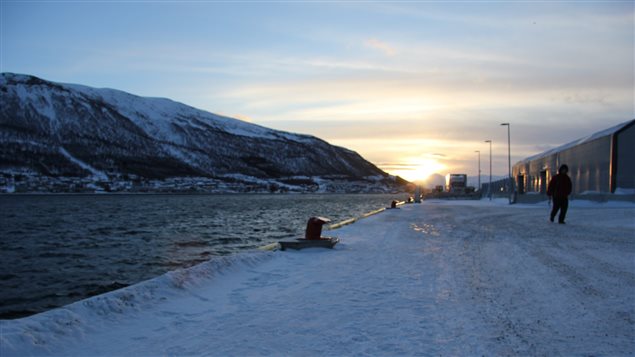 The Arctic port of Tromso, Norway is shown as the sun goes down. Snow-covered mountains are on the left and a man walking along the port is pictured on the right in a dark winter parka.