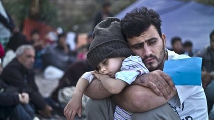 A Syrian refugee and his child wait at a resting point to board a bus on the Greek island of Lesbos in October. We see a dark-haired man with a slight, dark beard dressed in white. His enormous arms hug a child, who appears to be two or three, in his lap. The child wears a grey toque. He is sound asleep.