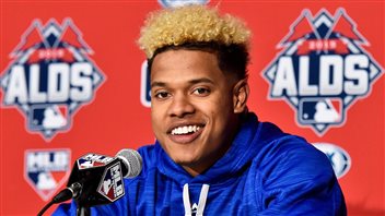  Marcus Stroman, a short man (he stands 1.73 metres) with enormous will and confidence, gets the start for the Blue Jays at their home opener on Friday night at Rogers Centre. We see a handsome young black man sitting behind a microphone. His hair on top has been dyed blond. (The sides of head display dark hair.) He wears a dark blue draw-string sweatshirt and a wide, slightly crooked smile, displaying a set of very white teeth. Behind him is a wall logo with the letters ALDS (for American League Division Series).