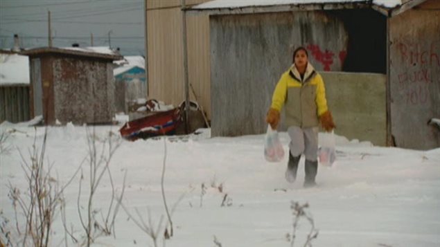 Extreme poverty at the Attawapiskat First Nation has led to over 100 suicide attempts. We see a girl of about 15 carrying two shopping bags walking through snow. Behind her are run-down buildings and shacks. She has beautiful dark skin and black hair and wears at yellow and tan winter jacket above grey pants and winter boots.