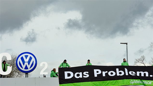 Volkswagen is continuing its efforts to weather its emission-rigging storm. Under dark grey skies, we see five people dressed in green windbreakers holding up a long black sign that says "Das Problem" printed in white block letters. They appear to be atop a Volkswagen facility of some sort as the company's round blue-and-white logo stands at the left of the photo.
