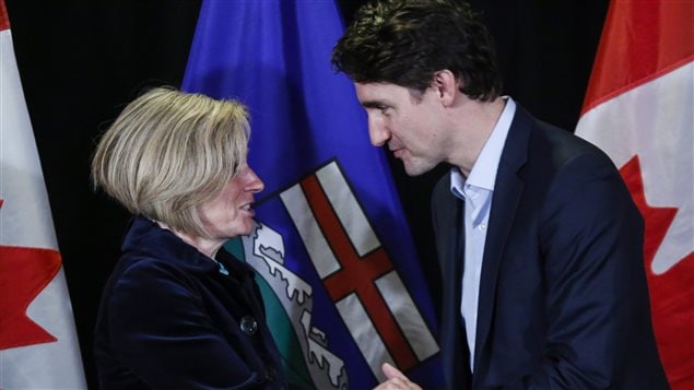 Prime Minister Trudeau, right, met with Alberta Premier Rachel Notley following meetings at a Liberal Party cabinet retreat in Kananaskis, Alta. on Sunday.  Ms. Notley is at the left of the photo, Mr. Trudeau on the right. They are engaged in a clasped hands shake with the taller Trudeau leaning in and looking the blonde-haired Notley directly in the eyes. She appears slightly taken aback. Both are dressed in blue jackets. The blue, white and red Alberta flag stands behind them .