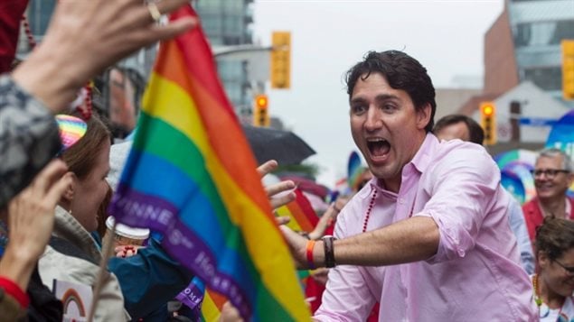 Prime Minister Trudeau (seen at last year's Gay Pride Parade in Toronto) wants his government to protect transgender rights. We see Trudeau in a dress shirt with the sleeves rolled up and his mount agape reaching for a blue, green, yellow and red flag being waved by a participant in the parade.