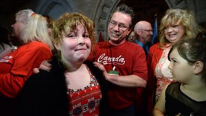  Charlie Lowthian-Rickert is comforted by her dad, Chris Rickert, her mom, Anne Lowthian, right, and friends following Justice Minister Jody Wilson-Raybould's launch of the transgender rights bill. Chris Rickert wears a red tee-shirt with the word Canada written in script across the chest. He wears glasses and a smile as he places both his hands on Charlie's shoulders. Looking toward the camera, dressed in a dark sweater and red dress, she wears a look of great pride on her face. Anne Lowthian stands to the right of the picture. She is a large woman with blond hair whose face is a mix tears and laughter.  