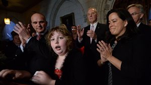 Charlie Lowthian-Rickert speaks as Justice Minister Wilson-Raybould, right, applauds on Parliament Hill on Tuesday. We see a young girl with blond, curly hair standing at the podium. As any 10-year-old might, Charlie has just a touch of bewilderment in her eyes as she speaks. Behind her to her left the justice minister, dressed in a black sweater, is applauding with a look of pride on her face. Behind the two, are several men, likely MPs, dressed in spiffy, dark suits.