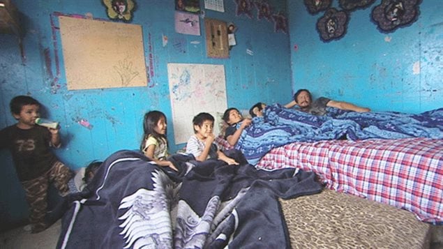  The First Nations community of Pikangikum in northwestern Ontario is one of many facing problems of overcrowding and poverty. A Canadian Centre for Policy Alternatives report says indigenous children in Canada are more than twice as likely to live in poverty as non-aboriginal children. We see a family of four, young aboriginal children and their parents huddled under blankets under two mattresses in a bed room that has chipped aqua blue paint on its walls. The walls are decorated with a series of aboriginal decorations.