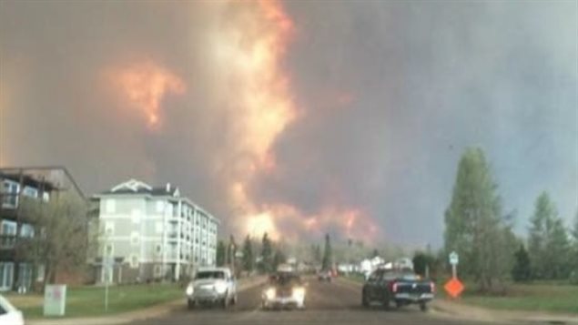 As the giant wildfire approached Fort McMurray on May 3, the first of the just under 90,000 people began their flight. Now, 8,000 others north of the city are being evacuated. We see two cars heading toward the camera. To their left, a black pickup truck heads the other way. In the background, we see great flames billowing into a charred sky.